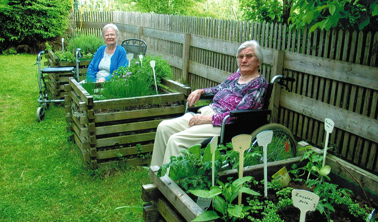 Zwei Senioren sitzen in geschütztem Garten zwischen Kräuterbeeten
